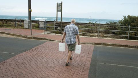 A man walks across the road with two water tanks after collect drinking water from pipes fed by an underground spring, in St. James, about 25km from the city centre, on January 19, 2018, in Cape Town.
Cape Town will next month slash its individual daily water consumption limit by 40 percent to 50 litres, the mayor said on January 18, as the city battles its worst drought in a century. / AFP PHOTO / RODGER BOSCH