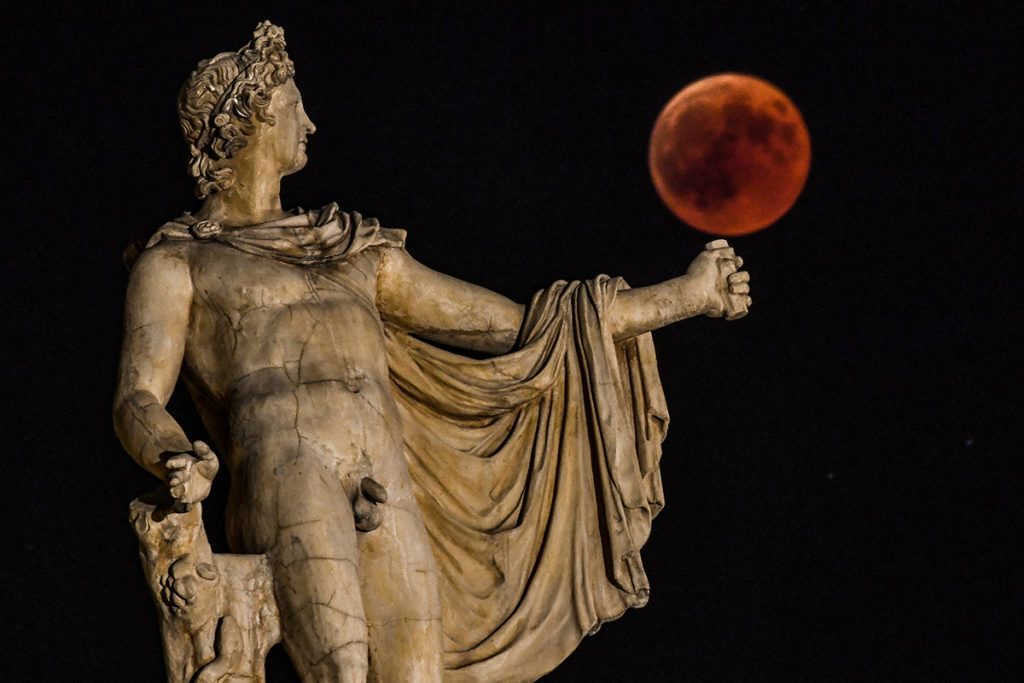 A picture shows the full moon during a "blood moon" eclipse beside a statue of the ancient Greek god Apollo  in central Athens on July 27, 2018. The longest "blood moon" eclipse this century began on July 27, coinciding with Mars' closest approach in 15 years to treat skygazers across the globe to a thrilling celestial spectacle. / AFP PHOTO / Aris MESSINIS